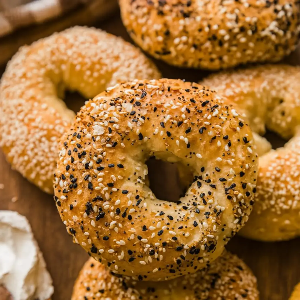 fresh-protein-sourdough-everything-bagels-baked-crust-sourdoughcravings Close-up of golden brown protein sourdough everything bagels topped with sesame, poppy seeds, garlic, and onion flakes on a rustic wooden surface