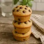 High Protein Sourdough Muffins That Fuel Your Day Naturally 3 Three high protein sourdough muffins stacked on a rustic wooden surface with chocolate chips, a checkered napkin, and blurred basil in the background.