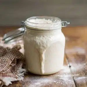 How to make a sourdough starter from scratch (no fail, beginner guide) – active bubbly starter in a glass jar on rustic wood with brown-and-white gingham tea towel
