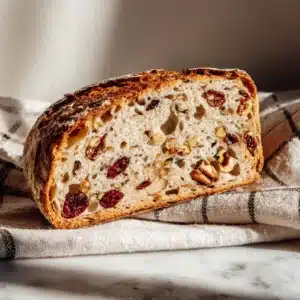Close-up of a sliced sourdough loaf filled with cranberries, pecans, and seeds on a checkered cloth.