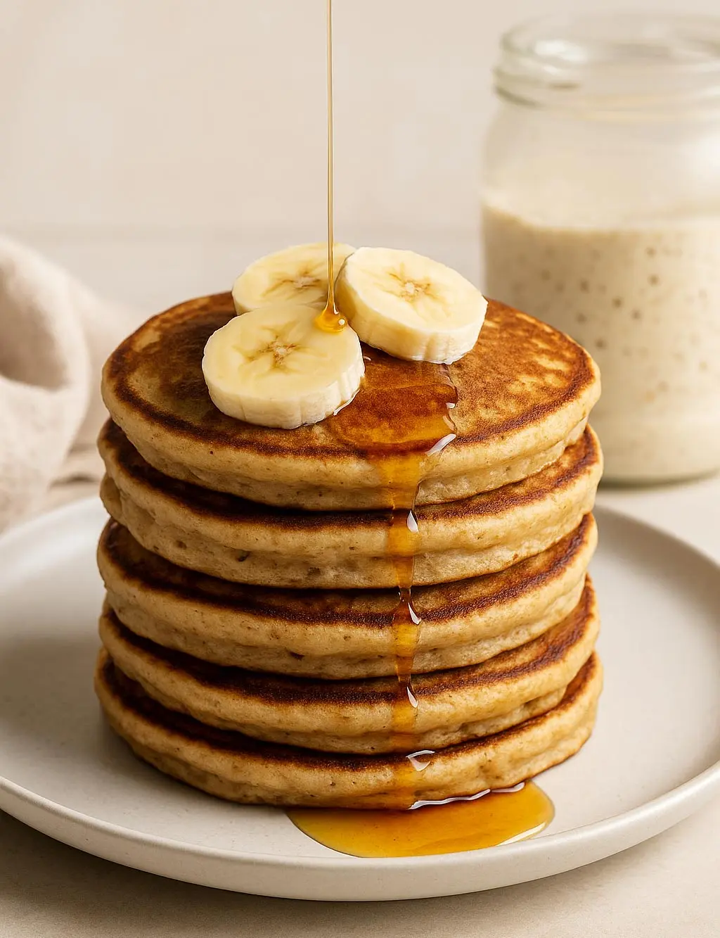 sourdough-protein-pancakes-banana-syrup Stack of protein sourdough pancakes topped with banana slices and drizzled with syrup